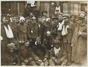 British and Indian wounded at Neuve Chapelle, on the way to the hospital base, 1915. One of the British is wearing a German helmet. Shows a group of wounded Germans, British and Indians next to a hospital train. (National Army Museum)